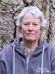 Portrait of a Jane Guiltinan, woman with short grey hair and a purple sweatshirt standing in front of tree so large it fills the frame.