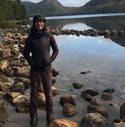 Smiling young woman in bicycle helmet standing in creekbed with green mountains in the background.