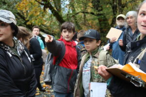 Schoolkids and adults on riverbank looking in direction of boy pointing