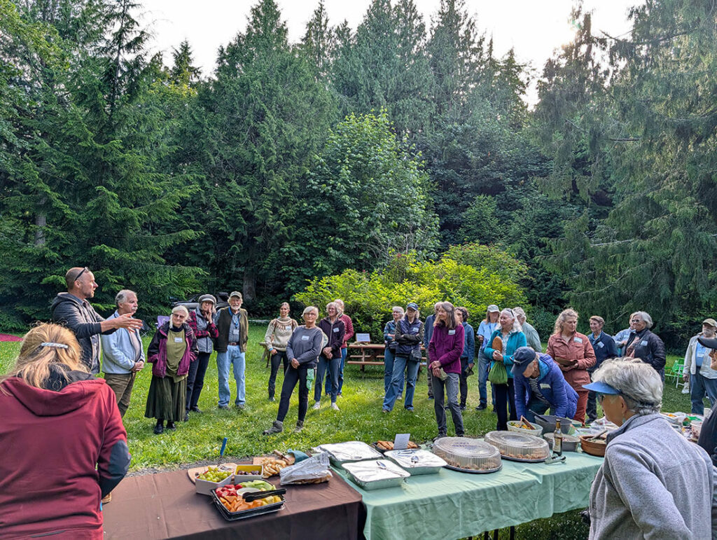 Group of 25 people beside food-laden picnic tables at outdoor park.