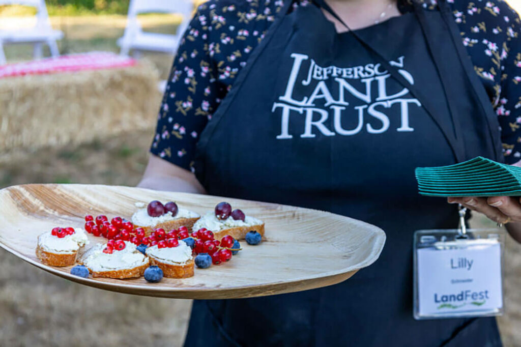 Close up of person in black apron reading "Jefferson Land Trust" holding tray of crostinis and red berries.