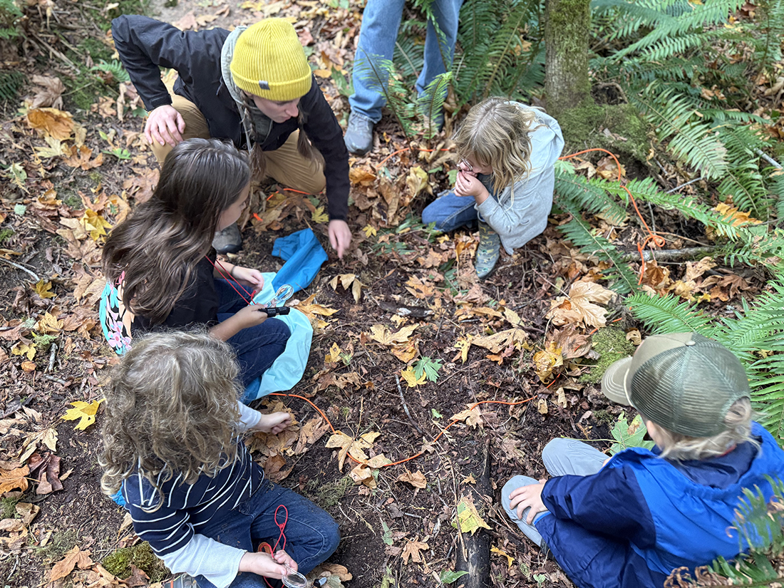 Group of young students and an adult sitting on the ground looking at something.