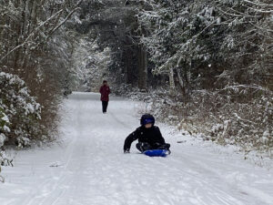 Photo of child sledding on a snowy trail in the Quimper Wildlife Corridor with an adult walking behind.