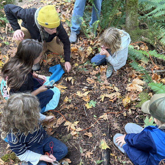 Group of young students and an adult sitting on the ground looking at something.